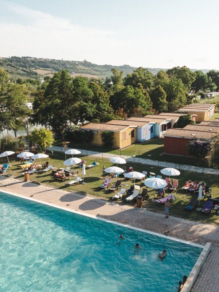 Piscina all'aperto con sdraio e ombrelloni, vista di colline e bungalow in lontananza.