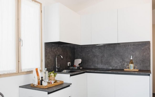 Cucina moderna con mobili bianchi e piano di lavoro scuro, provvista di alcuni utensili e bottiglie.