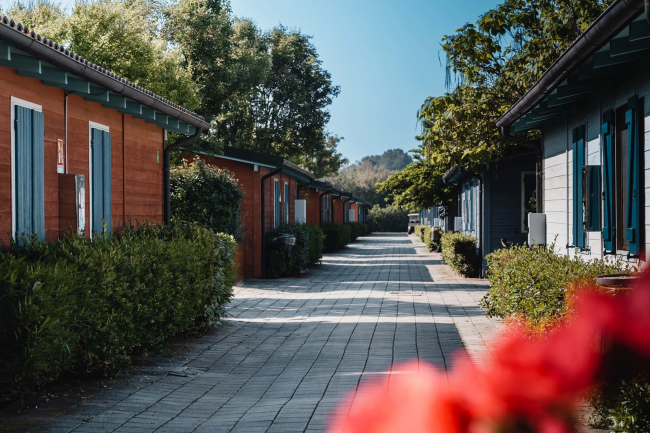 Un viale di bungalow colorati con finestre blu, contornato da siepi e alberi verdi.