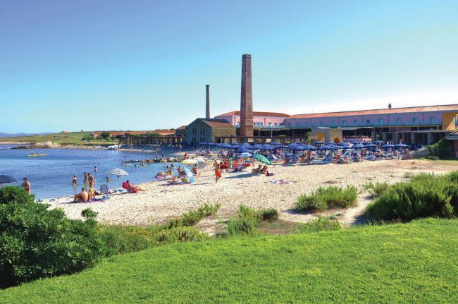 Spiaggia affollata con ombrelloni e edificio con ciminiere.