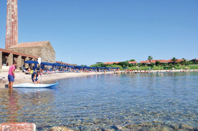 Spiaggia con mare cristallino, ombrelloni e un vecchio edificio in pietra.
