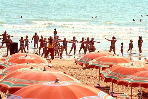Persone partecipano a un'attività di gruppo sulla spiaggia, con ombrelloni colorati in primo piano.