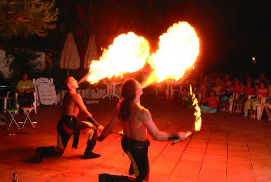 Due artisti di strada sputano fuoco davanti a uno spettacolo serale con pubblico seduto.