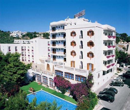 Edificio bianco con piscina e fiori fucsia, circondato da auto parcheggiate.