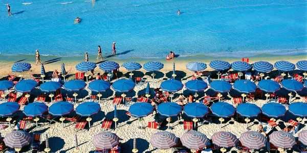 Spiaggia con numerosi ombrelloni blu e persone che camminano sul bagnasciuga del mare azzurro.