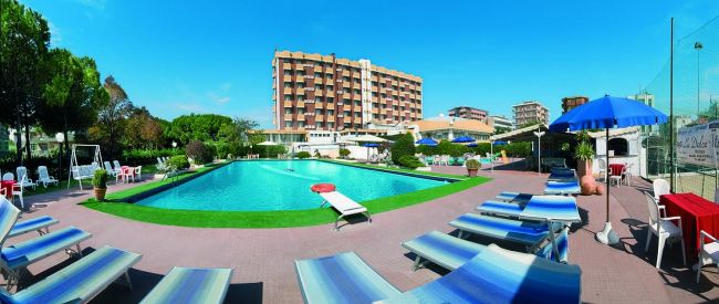 Piscina all'aperto con sdraio e ombrelloni blu, edificio sullo sfondo sotto un cielo azzurro.