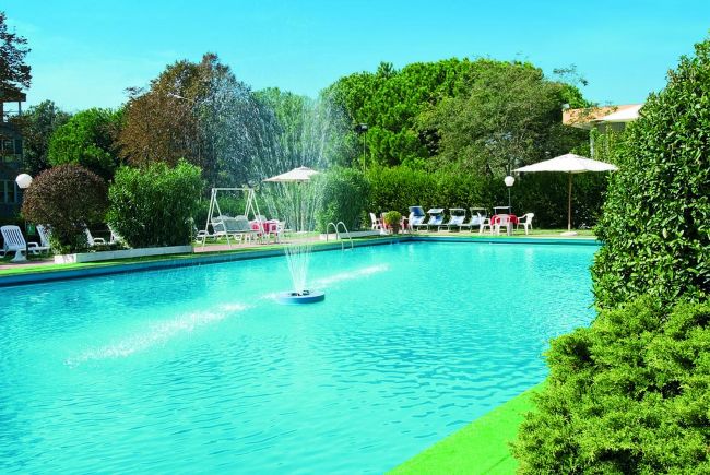 Piscina con fontana centrale, circondata da vegetazione e lettini bianchi sotto ombrelloni.