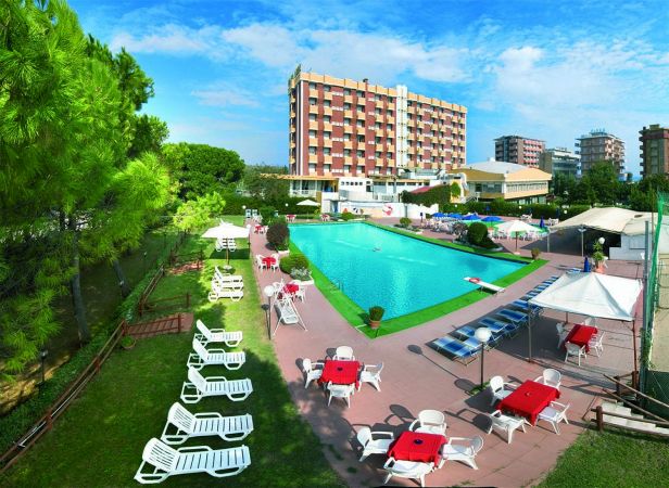 Piscina con sdraio e tavoli rossi, circondata da giardino e grandi edifici sullo sfondo.