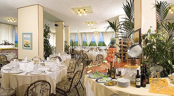 Sala da pranzo elegante con tavoli apparecchiati, piante decorative e buffet di cibo e vino.