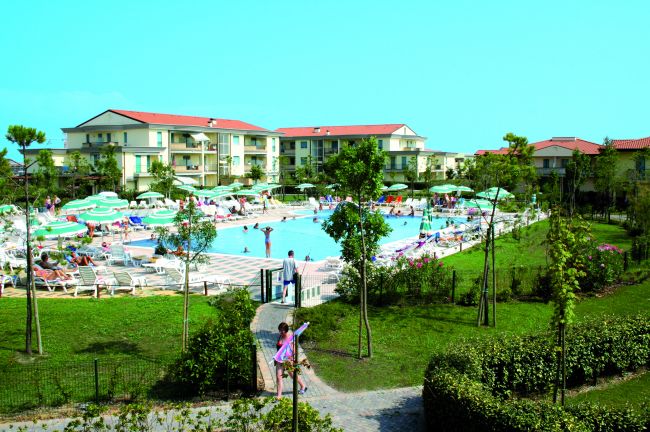 Piscina affollata con lettini e ombrelloni, circondata da prati e edifici residenziali.