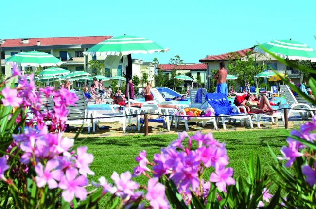 Persone rilassate su sdraio sotto ombrelloni verdi, intorno a una piscina.