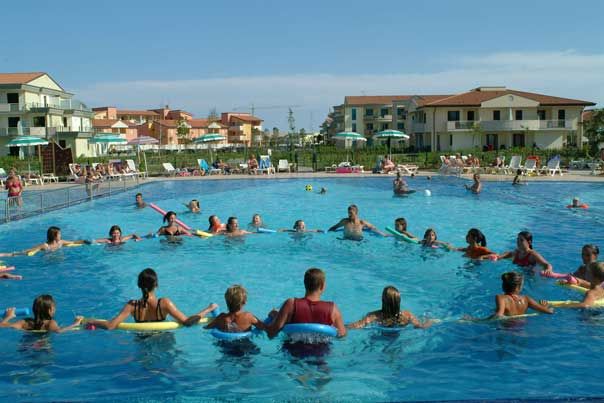 Gruppo di persone in cerchio in una grande piscina, con edifici residenziali sullo sfondo.