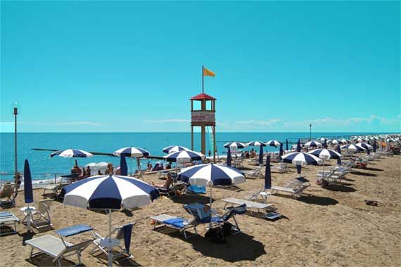 Spiaggia con file di ombrelloni blu e bianchi, sdraio e una torretta di salvataggio vicino al mare.