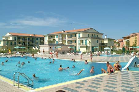 Una piscina all'aperto con persone che nuotano, circondata da edifici a due piani e ombrelloni.