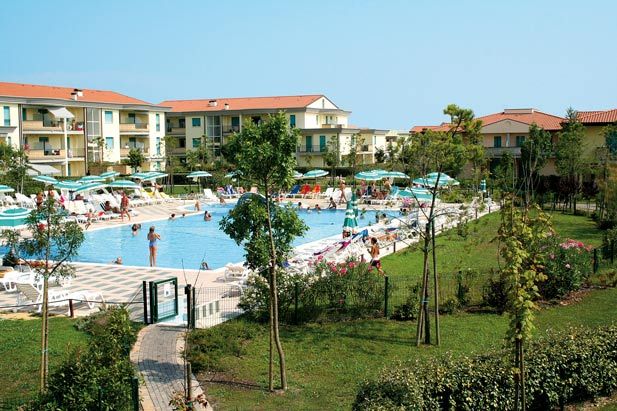 Vista di una piscina con lettini, ombrelloni, edifici circostanti e un giardino alberato.