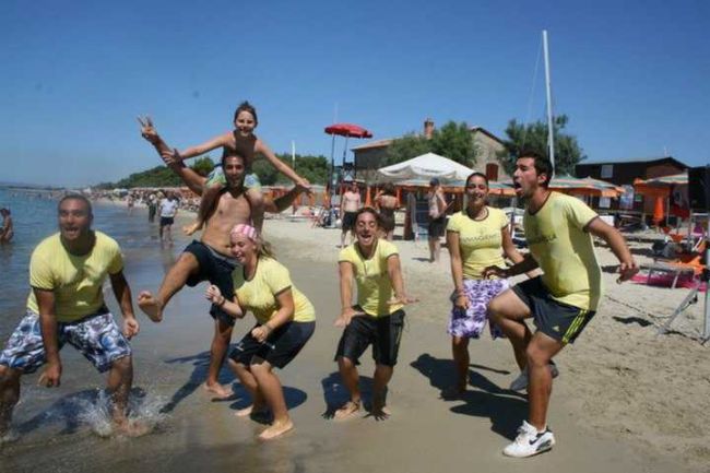 Un gruppo di persone in magliette gialle che sorride e fa pose divertenti su una spiaggia.
