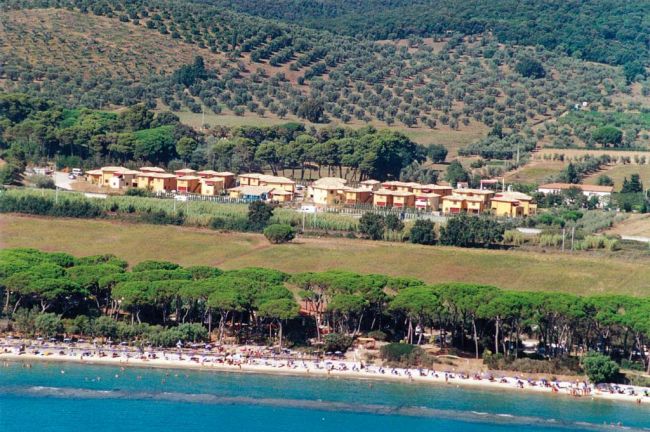 Complesso residenziale vicino a una spiaggia con pini e colline sullo sfondo.