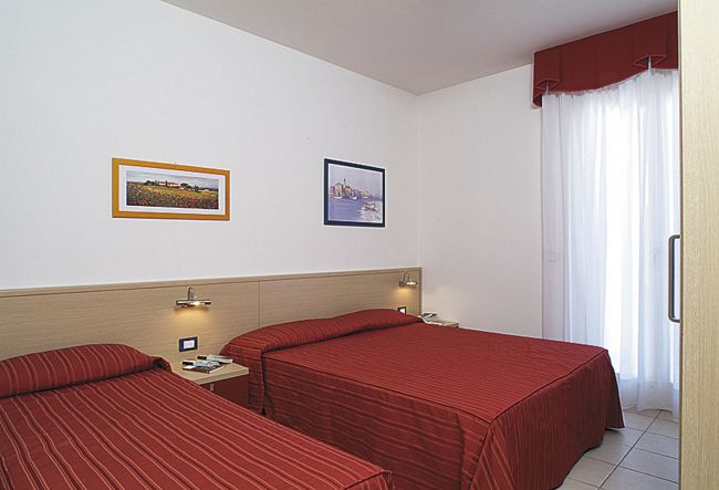 Camera con due letti singoli, copriletti rossi, due quadri alle pareti, e una tenda bianca.