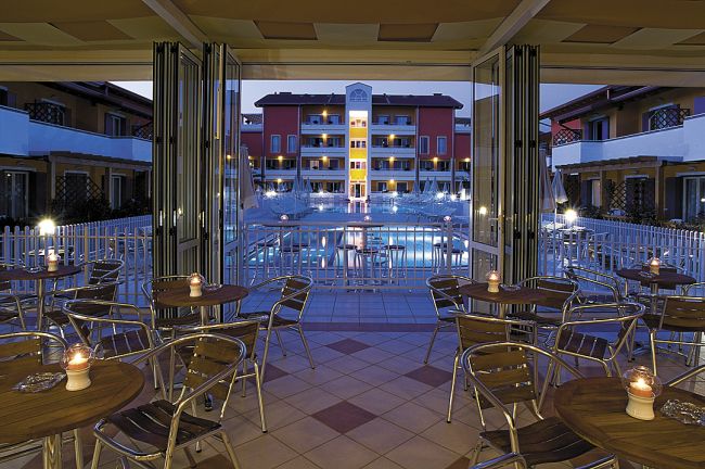 Terrazza con tavoli e sedie, vista serale su piscina e edificio illuminato.
