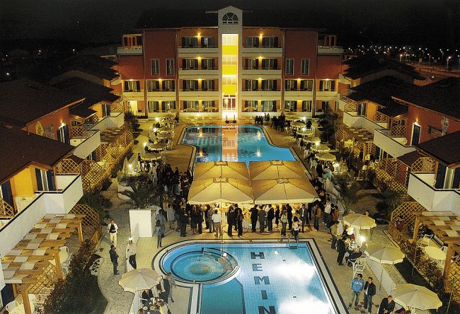 Vista notturna di un hotel con piscina illuminata e ospiti attorno a tavoli e ombrelloni.