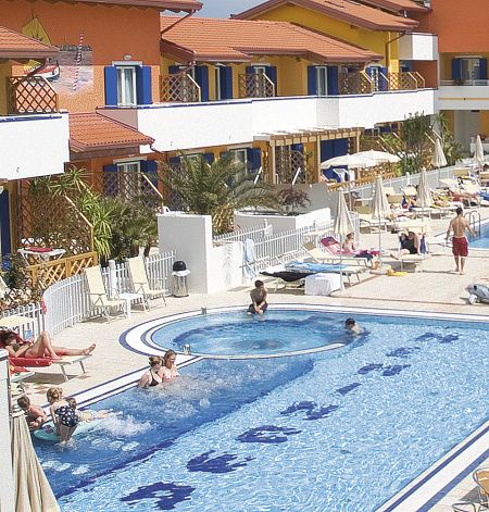 Piscina all'aperto con ospiti che nuotano, lettini e ombrelloni, di fronte a edifici colorati.