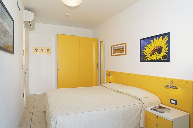 Camera con letto matrimoniale, pareti bianche, decorata con quadri e dettagli gialli.