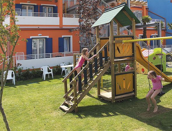 Parco giochi con scivolo e casetta in legno, bambini che giocano. Sfondo con edificio arancione.