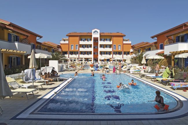 Piscina con vari nuotatori e sdraio intorno. Edifici arancioni e cielo blu sullo sfondo.