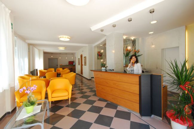 Reception di un hotel con sedie gialle e personale alla scrivania. Piastrelle pavimentali a scacchi.