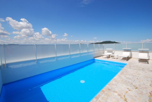 Una piscina azzurra su una terrazza con lettini bianchi e un cielo blu con alcune nuvole.