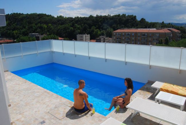 Due persone sedute sul bordo di una piscina su una terrazza con vista su colline e edifici.
