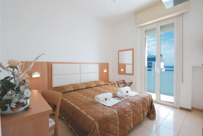 Camera d'albergo con letto matrimoniale, pareti bianche, balcone e vista mare.