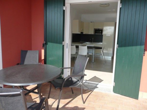 Terrazza con tavolo e sedie nere. Portafinestra con persiane verdi che conduce a una cucina moderna.