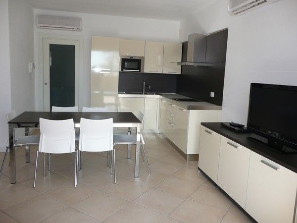 Cucina moderna con mobili chiari, tavolo nero con sedie bianche e TV su mobile.