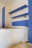 Letto con copriletto bianco, scaffali blu a muro, armadio blu e pavimento in legno chiaro.