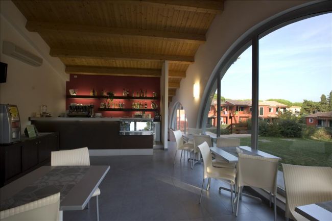 Interno di un bar moderno con ampie vetrate che offrono vista su un giardino.