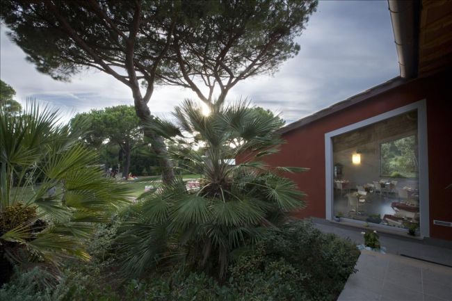 Un edificio rosso con ampia finestra accanto a palme e alberi sotto un cielo nuvoloso.