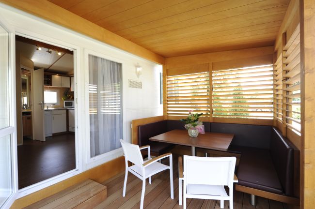 Veranda con tavolo in legno, sedie bianche e divani marroni. Interno di una cucina visibile.