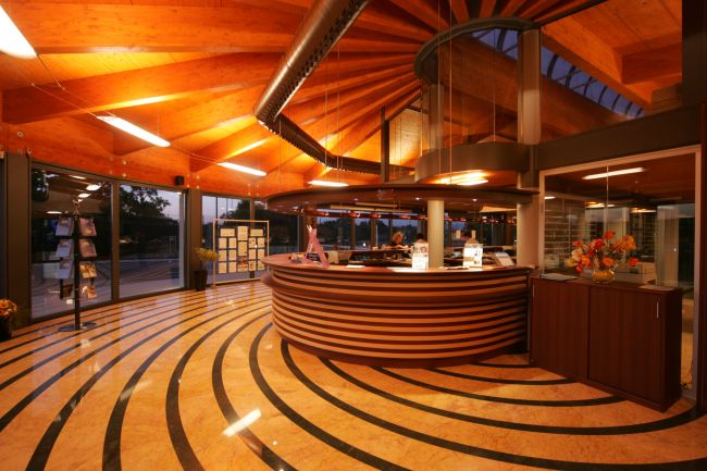 Hall con reception circolare, pavimento a strisce ondulate e soffitto in legno con luci calde.