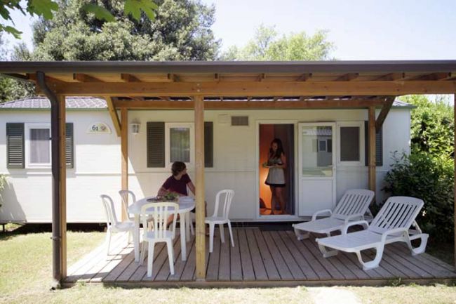 Esterno di una casa mobile con terrazza, tavolo e sedie da giardino sotto una tettoia.