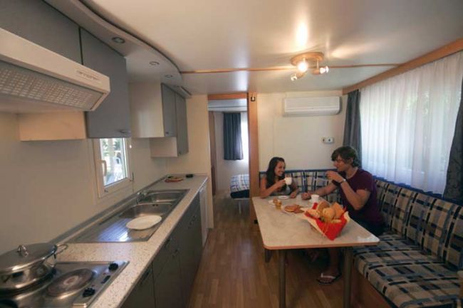 Interno di una roulotte con cucina e tavolo. Due persone mangiano insieme.