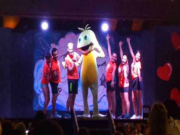 Personaggi sul palco con un costume giallo tra luci di scena.