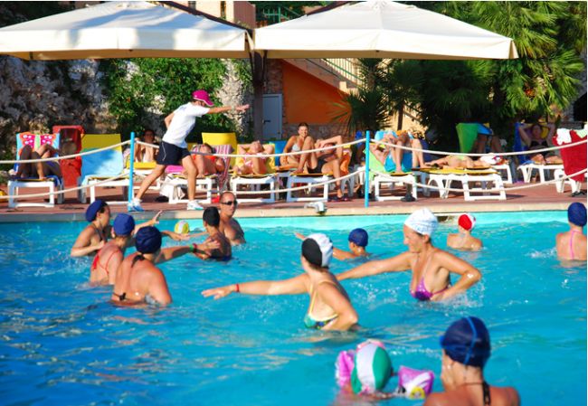 Persone che giocano in piscina, lettini e ombrelloni sullo sfondo.