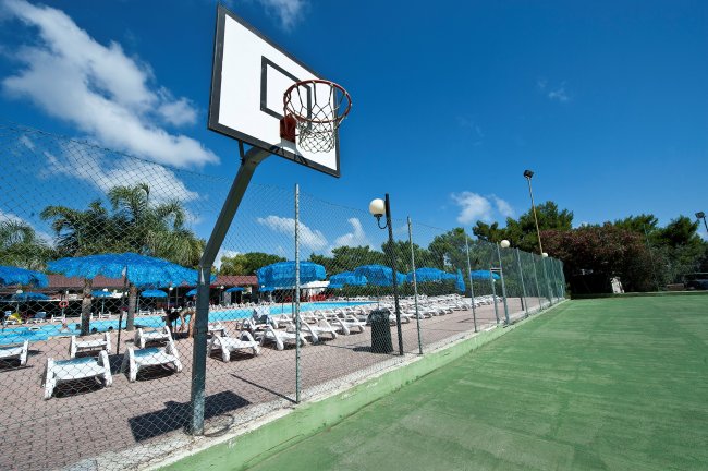 Campo da basket vicino a piscine con lettini e ombrelloni blu.