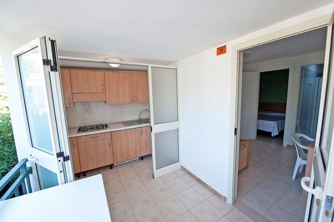 Cucina ad angolo con armadietti in legno e porta aperta verso una stanza.