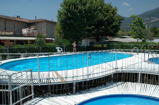 Piscina all'aperto con ponte bianco e area giochi, circondata da alberi e edifici.