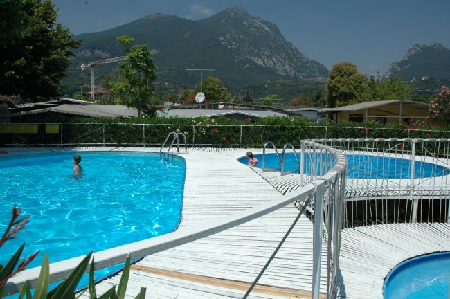 Due piscine con passerella bianca, montagna sullo sfondo. Persone nuotano.